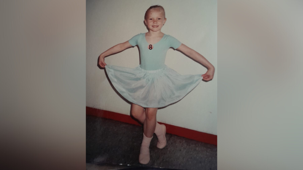 Melissa Hamilton as a child. She is wearing a light blue leotard with light blue shirt and pink sock. The number 8 is on her leotard. Her hair is slicked back and she is holding her shirt up at the sides and her right leg is bend backwards in a curtsey position.