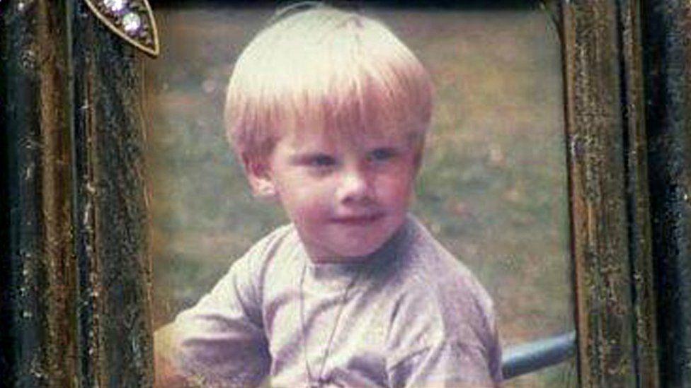 Colin Smith a little boy with blonde hair pictured in framed family photo. He is outside, wearing a grey T-shirt