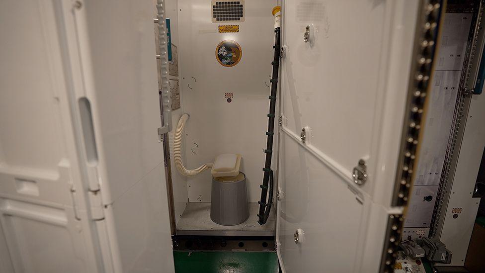 Toilet at Johnson Space Centre where astronauts train. The door is open to a small cubicle revealing a suction powered space station toilet. On the ground is a cylindrical grey base and on top of this is a white flat plastic oblong lid and seat. Coming out of this on the left hand side of the picture is a white corrugated pipe. On the right hand side is a black pipe with a yellow funnel at the end of it which is clipped to the wall. There is a sticker on the back wall of the cubicle that says international space station orbital outhouse team with a cartoon spaceman next to an old fashioned toilet.
