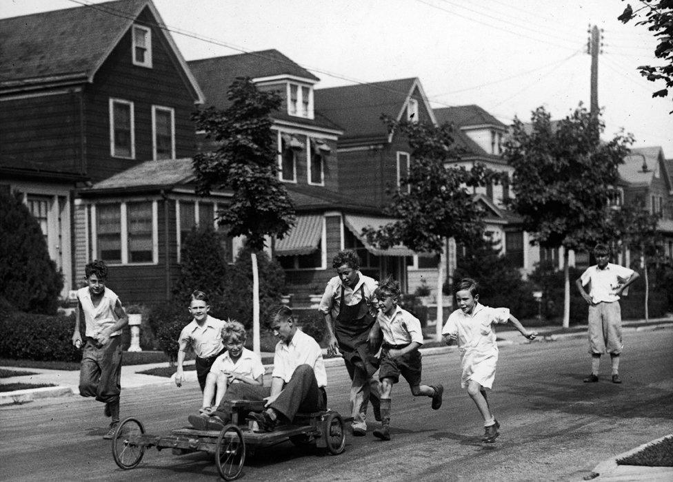 A group of boys on a residential street run after the go-kart that they have put together from an old motorcycle and a homemade chassis, 1930s.