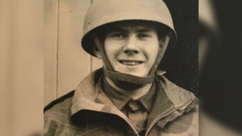 A sepia tone photo of Danny Gibbon at 17 years old in the army- wearing a camouflage jacket and rounded helmet with a chin strap