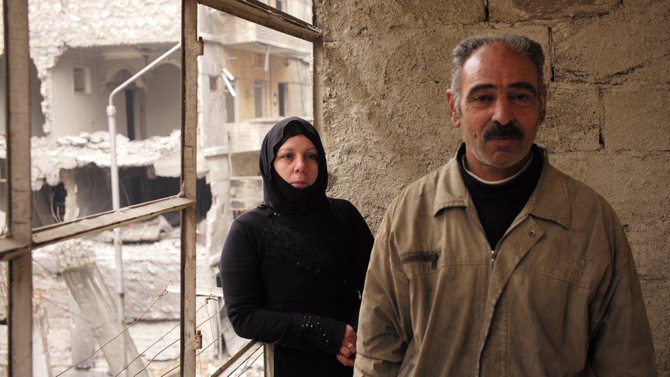 Abu Hussein and Umm Hussein stand next to blown out windows