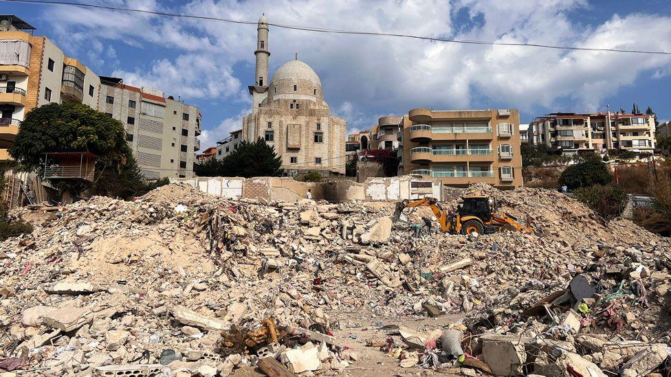 Rubble of the apartment block in the foreground, and in the background a few apartment blocks of various styles, flanking a mosque. A yellow digger picks through the detritus.