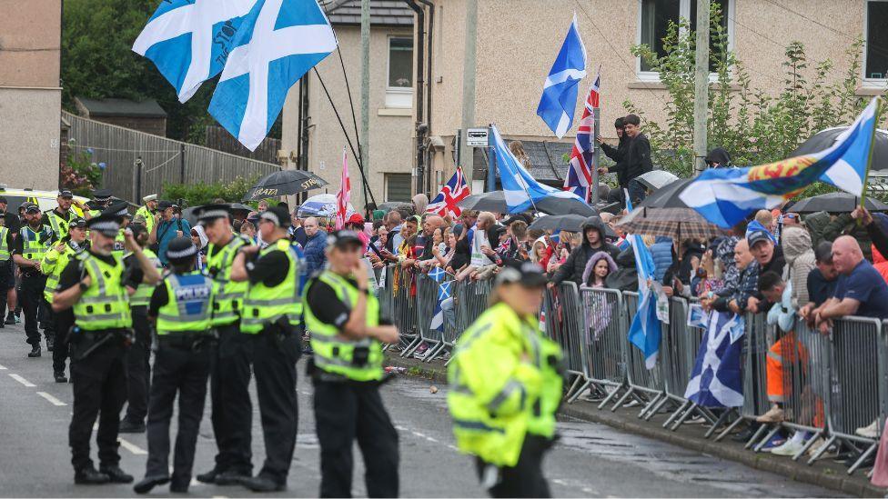 A line of police stand in front of protesters, who are behind metal barriers