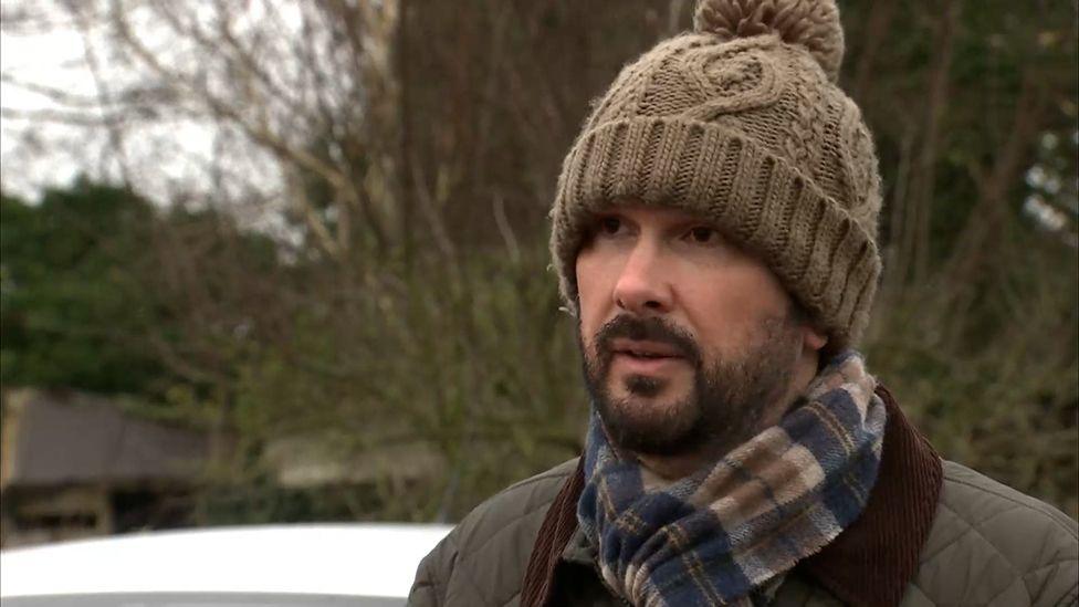 A bearded Paul Ansell standing by a river with trees in the background, in a warm coat, scarf and woolly hat