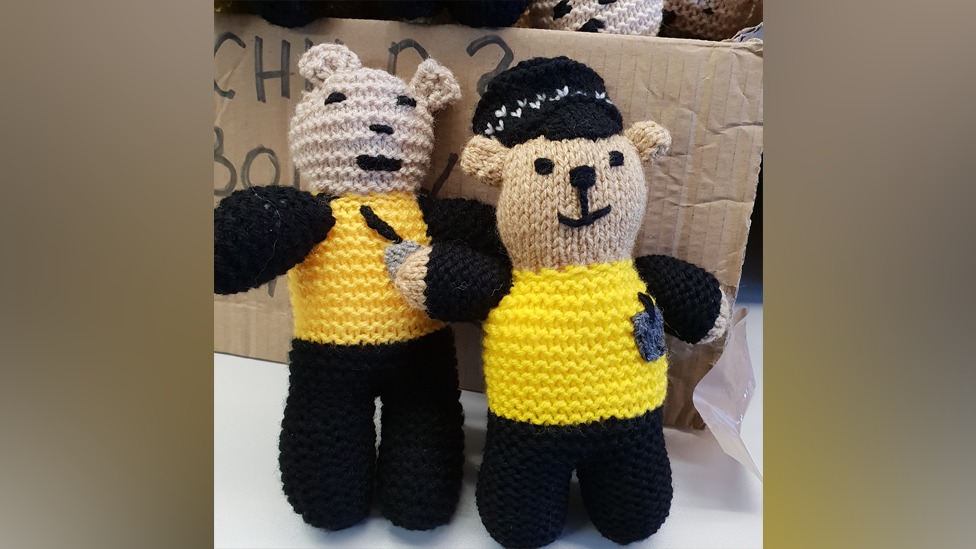 Two knitted teddies about the size of a hand. The bears are wearing a yellow and black police uniform and have little knitted police badges.