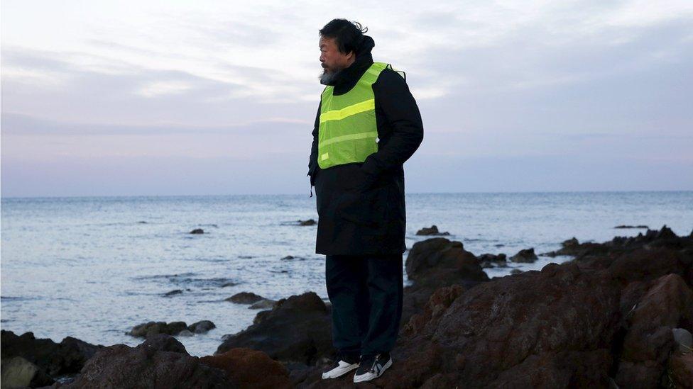 Ai Weiwei in a high vis jacket, standing on a rocky outcrop