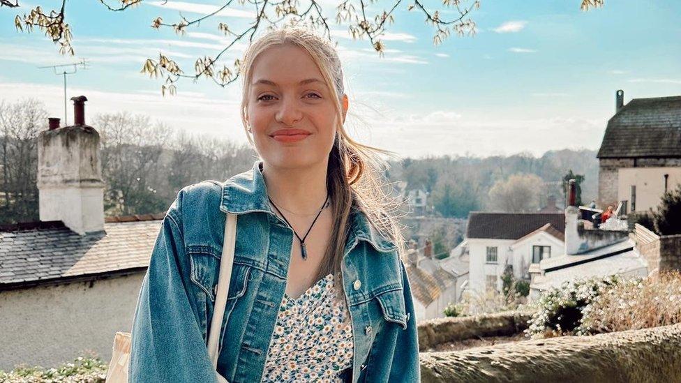 Jodie Powell is an 18-year-old white woman, she wears her long blonde hair tied back with her pony tail pulled over her left shoulder. She wears a denim jacket over a floral dress with a canvas tote bag over her right shoulder and a charm necklace on a black string. She is smiling at the camera and is photographed outside, with the roofs of houses and chimneys visible behind her as well as a line of trees against a pale blue sky