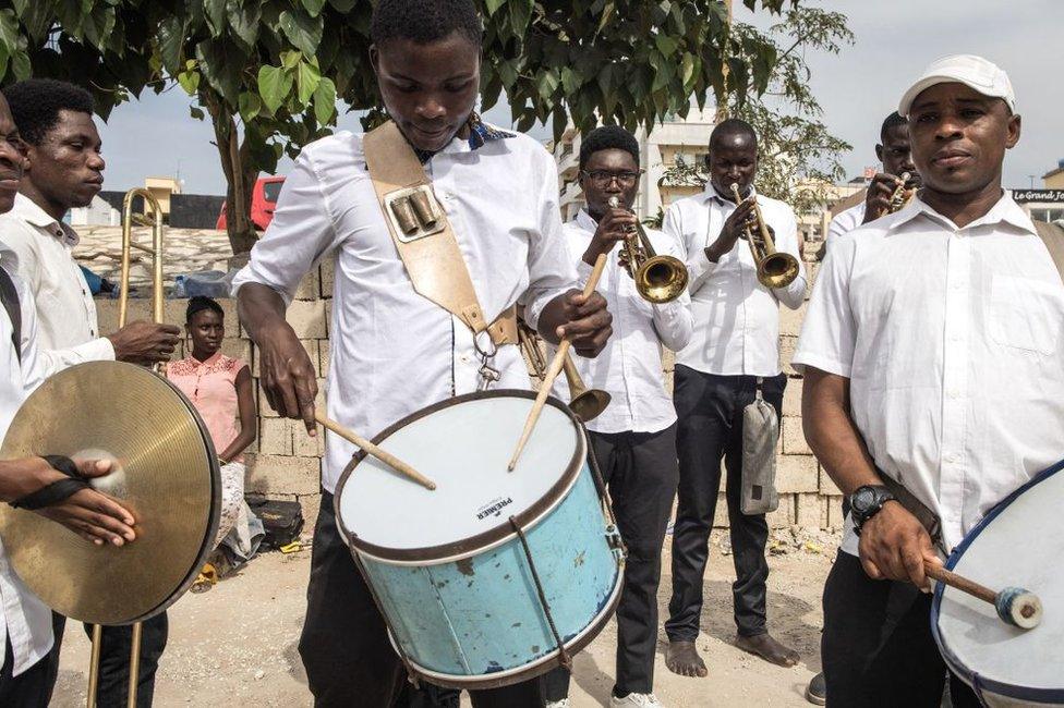 Men dressed in matching white shirts play trumpet, trombone, cymbals and snare drum.