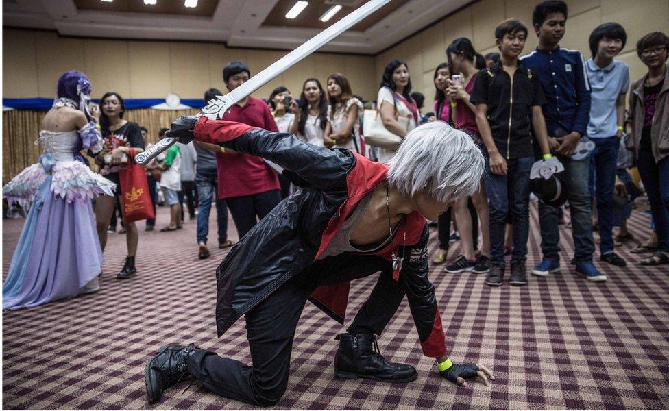 A man in a black and red cosplay outfit, carrying a plastic sword, crouches down on the floor in a pose in front of spectators at the event