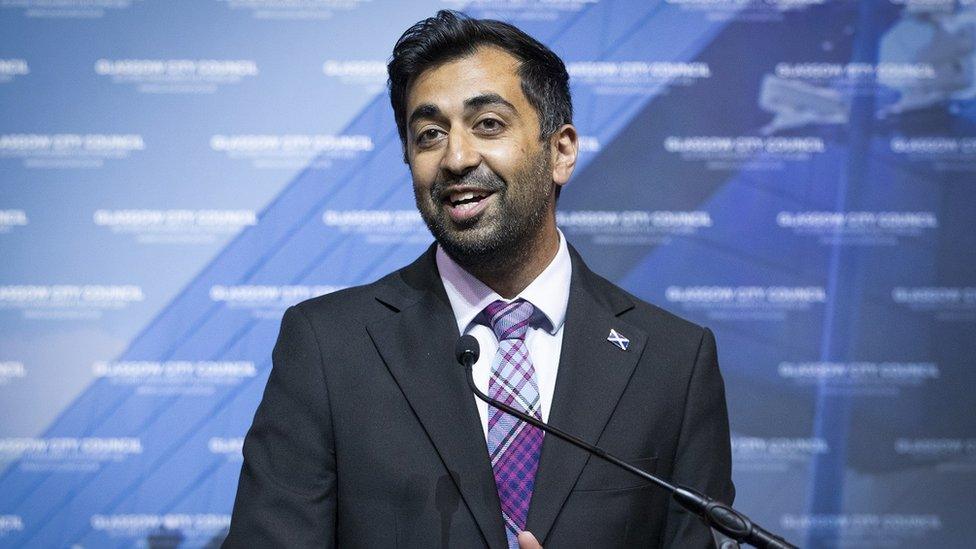 The SNP's Humza Yousaf stands in front of a microphone with a blue background in Glasgow