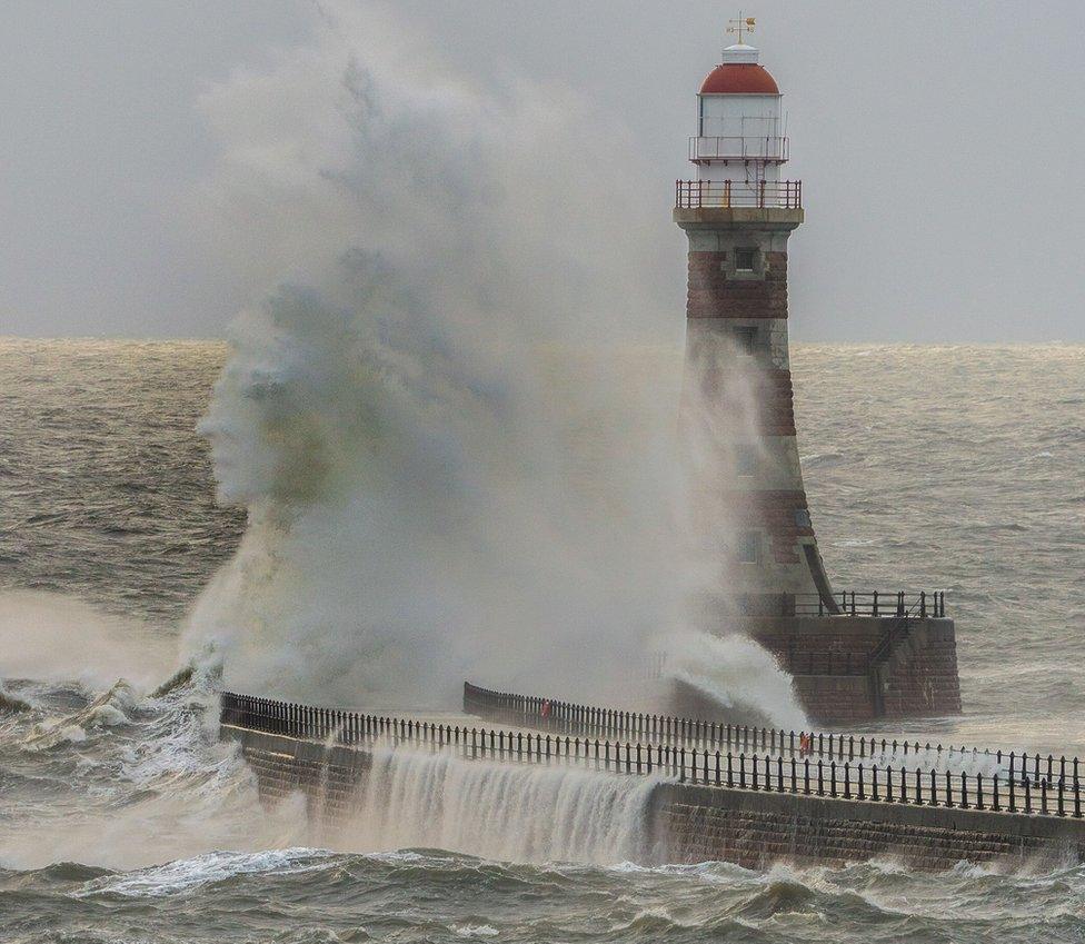 A massive wave breaks appearing in the shape of a human face next to a lighthouse