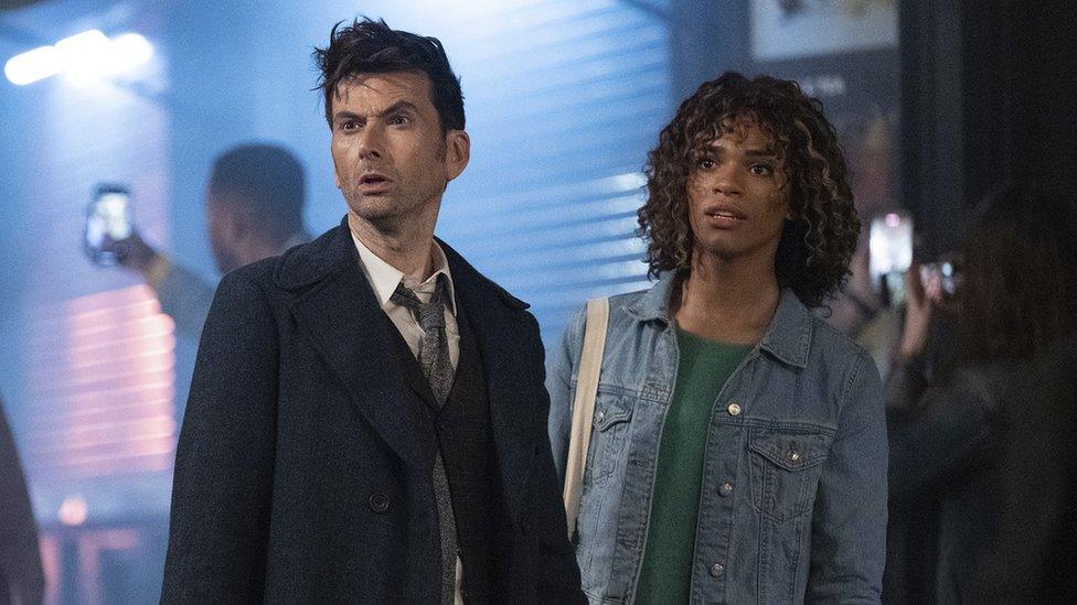 David Tennant as The Doctor and Yasmin Finney as Rose in Doctor Who. The Doctor has short messy brown air and wears a navy blue overcoat over a dark suit, white shirt and grey tie, tied loose. He has a serious expression with a furrowed brow and a short stubble. Rose, standing just behind him, has shoulder length curly brown hair. She wears a denim jacket over a green jumper and has a canvas tote bag over her right shoulder. Rose and the Doctor are seen outside on a street, with shutters and people taking photos on their phones behind them.