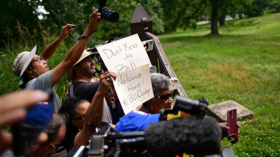 People hold signs outside the home of Bill Cosby after Pennsylvania's highest court overturned his sexual assault conviction and ordered him released from prison immediately