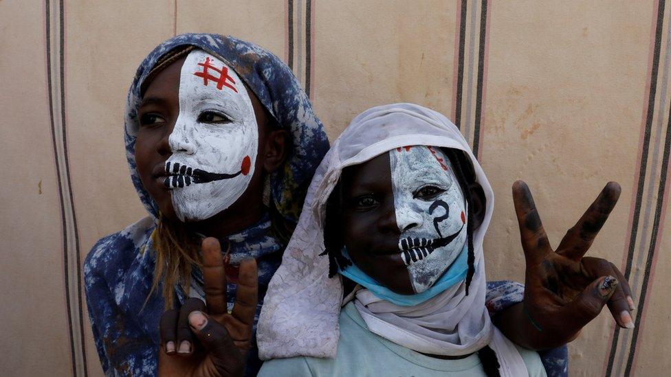 Sudanese girls with half painted faces make victory signs as they watch protesters demonstrating