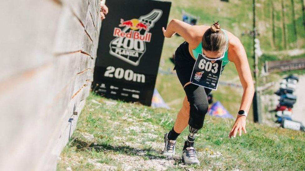 Milly Pickles at the 200m mark during the Red Bull 400 event in Slovenia. Millie, a white woman in her 20s, looks down as she strides up the incline. She has her brunette hair tied back in a bun and wears white framed sunglasses, a blue-green sports top and black shorts. She has a prosthetic limb on her right leg which is stretched in front of her left leg.