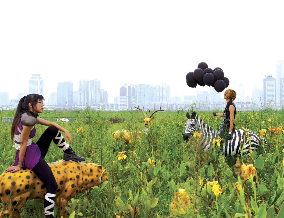 A photo by Cao Fei showing two people wearing costumes sitting on artificial animals with a city skyline in the background