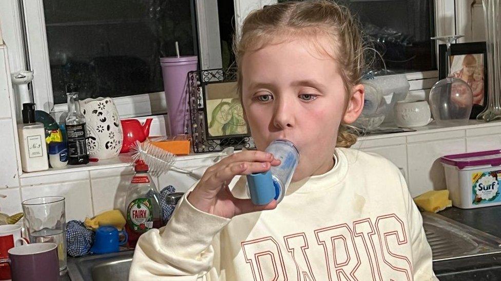 Girl with light-coloured hair breaths through an inhaler in a kitchen