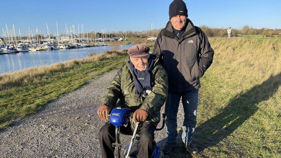 Elderly man in wheelchair wearing a cap and younger man with woolly hat behind beside a river