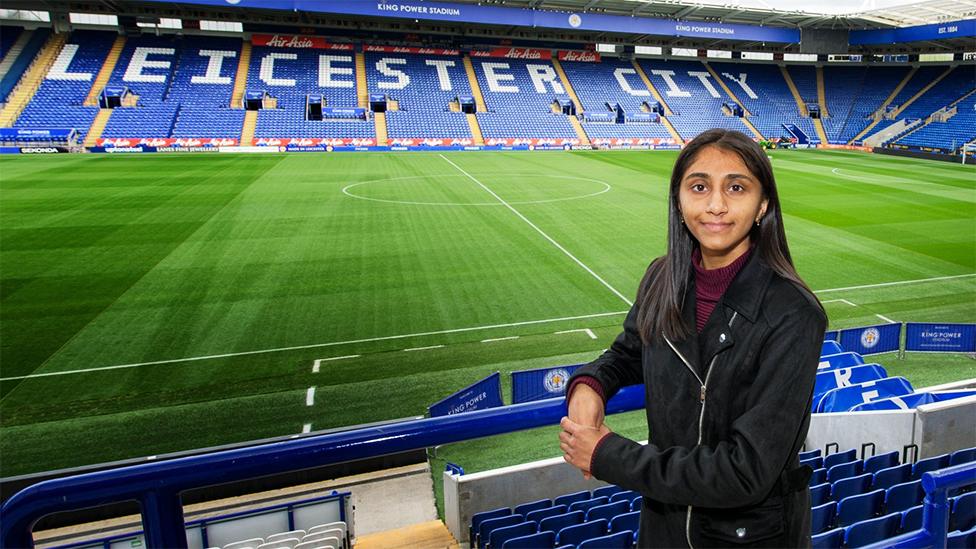 Renuka at Leicester City's stadium. Behind her is the green pitch and empty stands with blue seats, with white writing spelling out Leicester City. Renuka is looking at the camera, leaning on blue railings, with her left hand over her right hand. She is wearing a black jacket with a maroon turtle neck top.