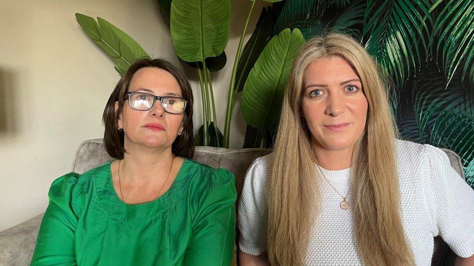 Two women sitting in front of a large plant. One has dark hair and glasses and wears a green top, the other has long light brown hair and wears a white top.