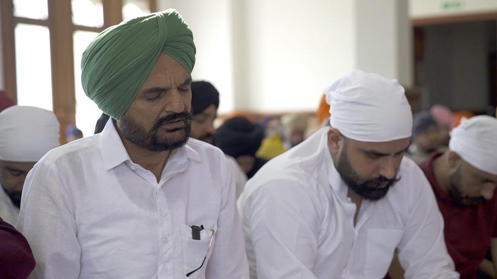 Balkaur Singh, wearing a green turban, sits on the left, while Sandy Joia sits on the right. Both men have their eyes closed in prayer. They are inside a Gurdwara, surrounded by other worshippers who are adopting a similar contemplative pose.