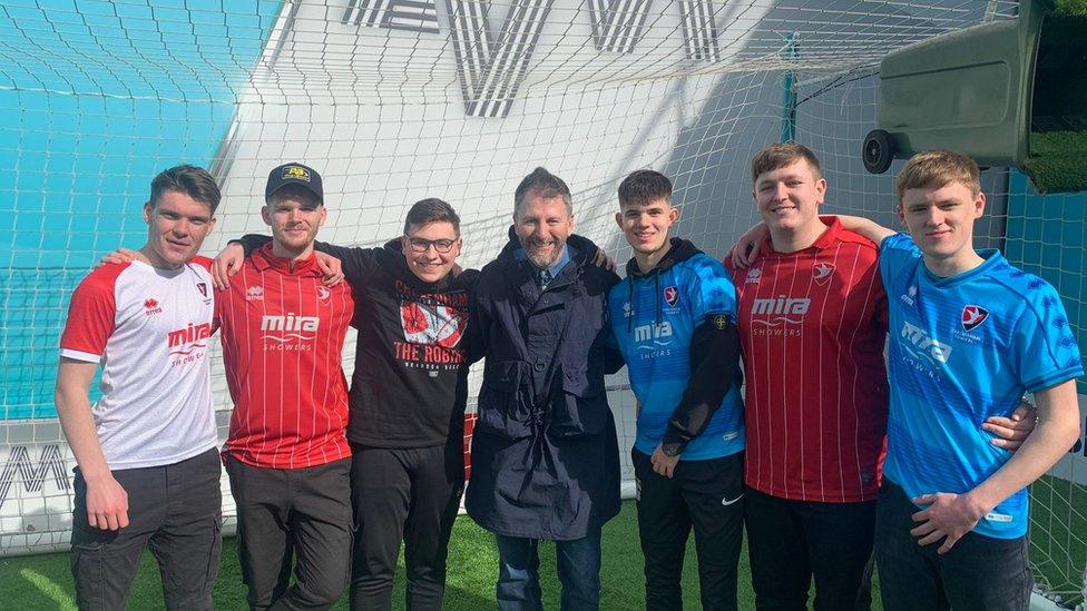 Fan Alex posing in front of the 'top bin' he scored a volley in, with fellow Cheltenham fans and presenter John 'Fenners' Fendley