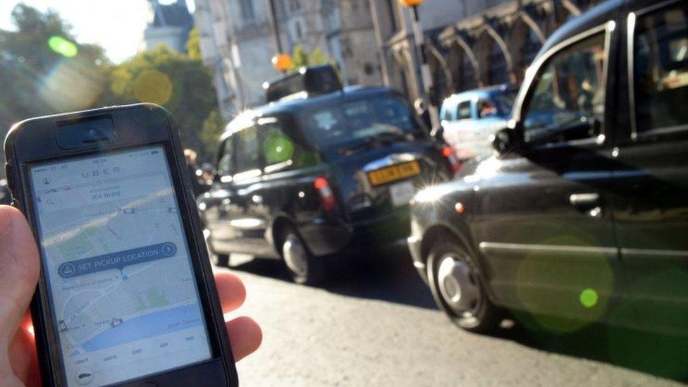 A smartphone displays the Uber app with black cabs visible in the background