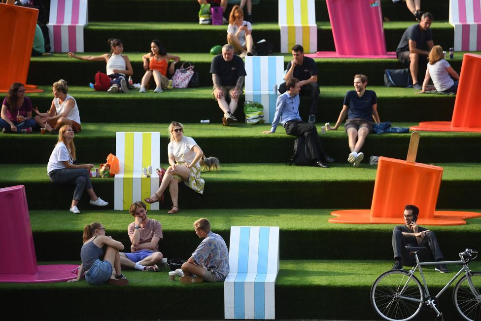 Groups of people sit on green steps outdoors, next to sculptures of melting ice lollies