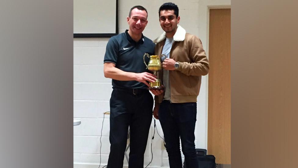 Rohan, an Asian man with Premier League referee Kevin Friend, a white man. The pair are holding either side of a gold trophy. Kevin is wearing a black top with a Nike brand in the corner and black trousers. Rohan is wearing a brown jacket with white fur at the top and a silver watch on his left hand.