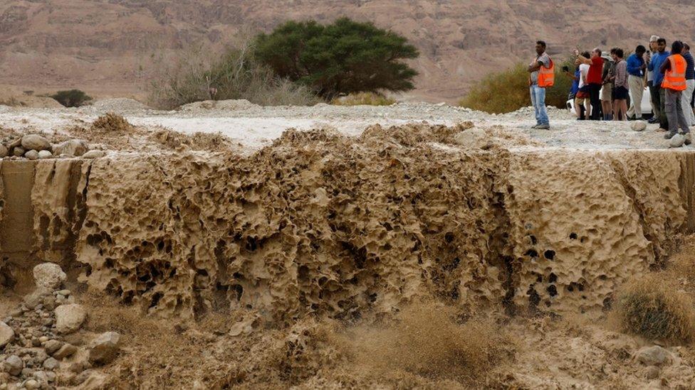 Israelis watch flooded water running through a valley, blocking the main road along the Dead Sea in the Judean desert