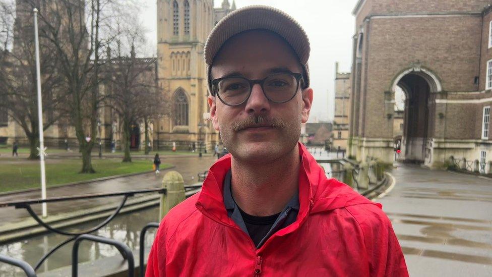 Sam Kidel in a red Acorn branded jacket and beige cap, wearing black thick-rimmed glasses