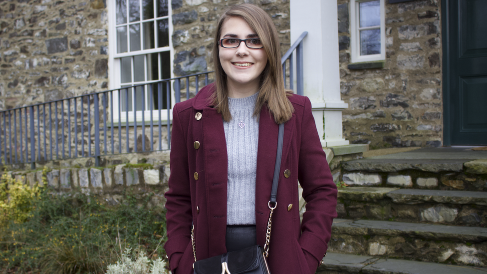 Elin wearing a coat and sweater standing in front of a building with traditional style stone wall