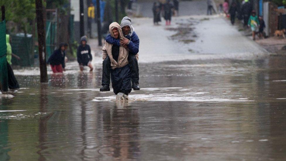 A man carries an elderly man across a flooded road during heavy flooding as a result of a storm in Sir Lowry's Village, close Somerset West on September 25, 2023.