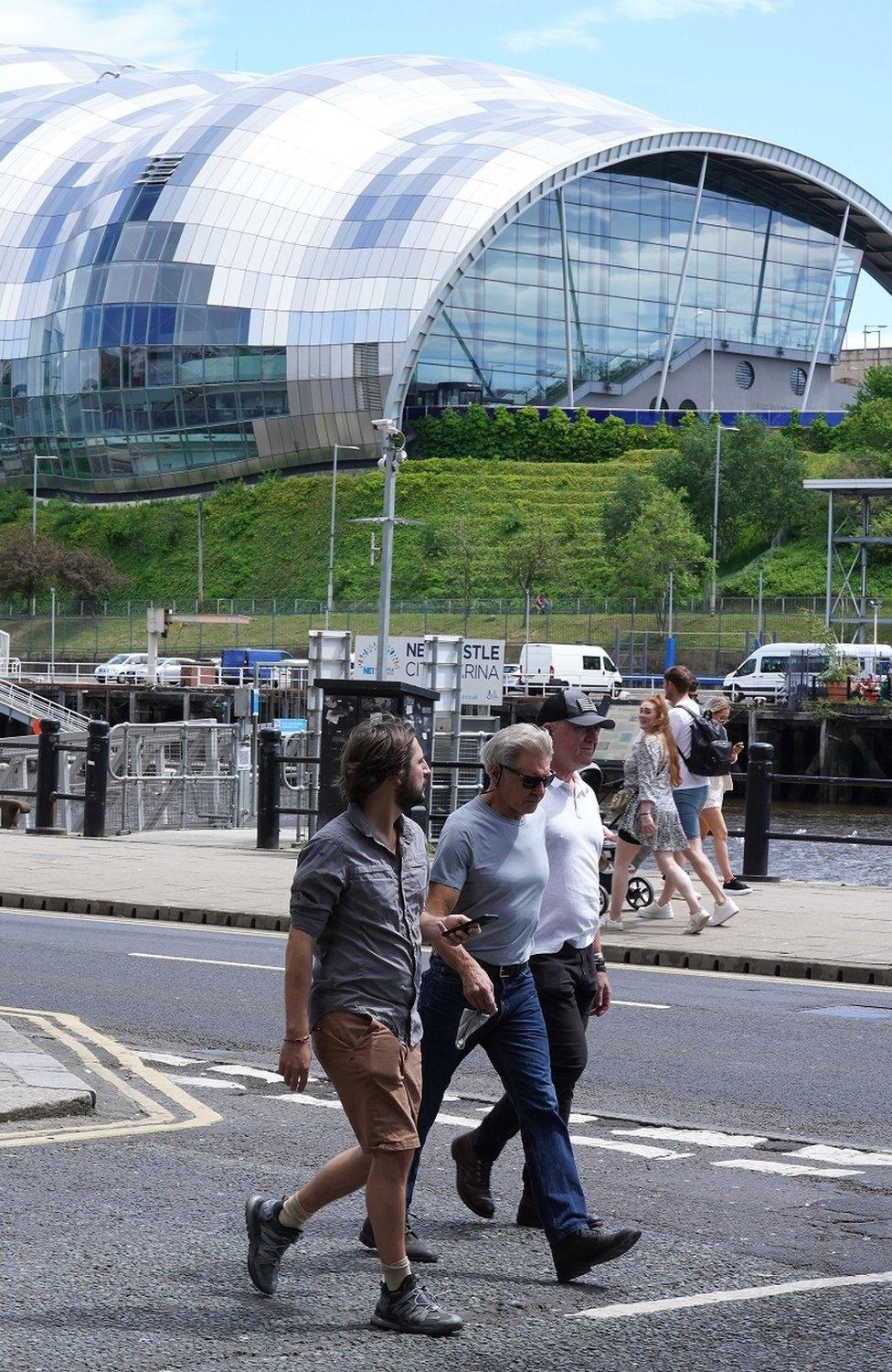 Harrison Ford walking along Newcastle Quayside, with two other men, with the Sage Gateshead in the background