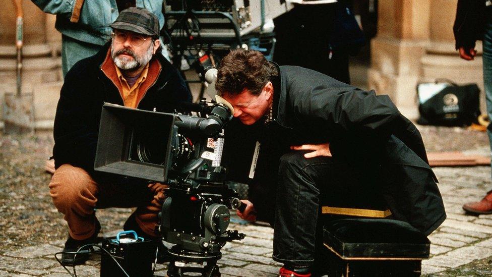 John Madden peers into the viewfinder of a large-format film camera on the ground, surrounded by other crew