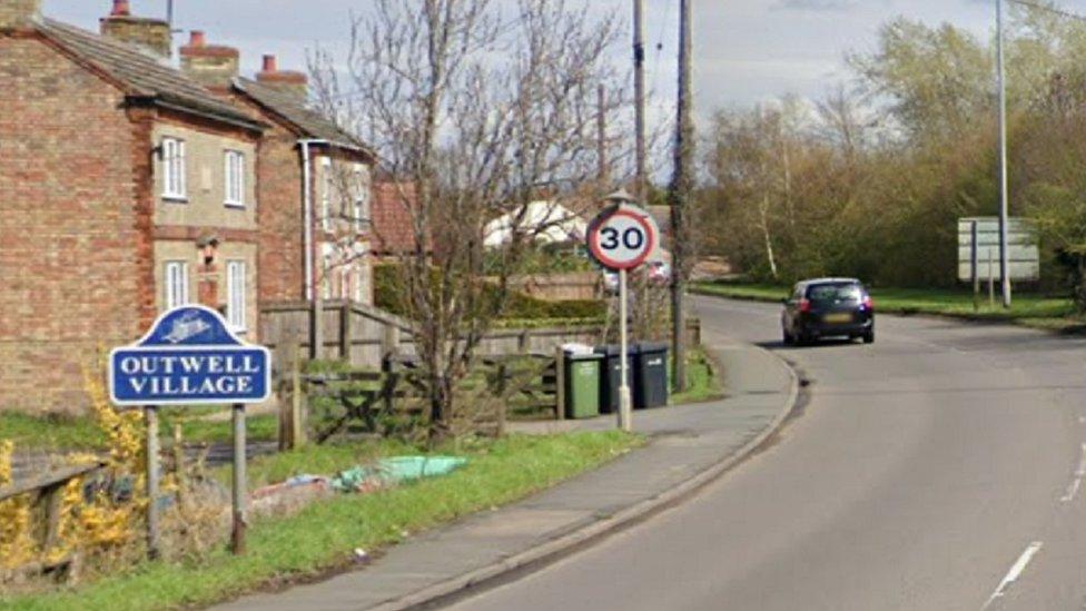 Main road into Outwell with blue village sign and houses to the left