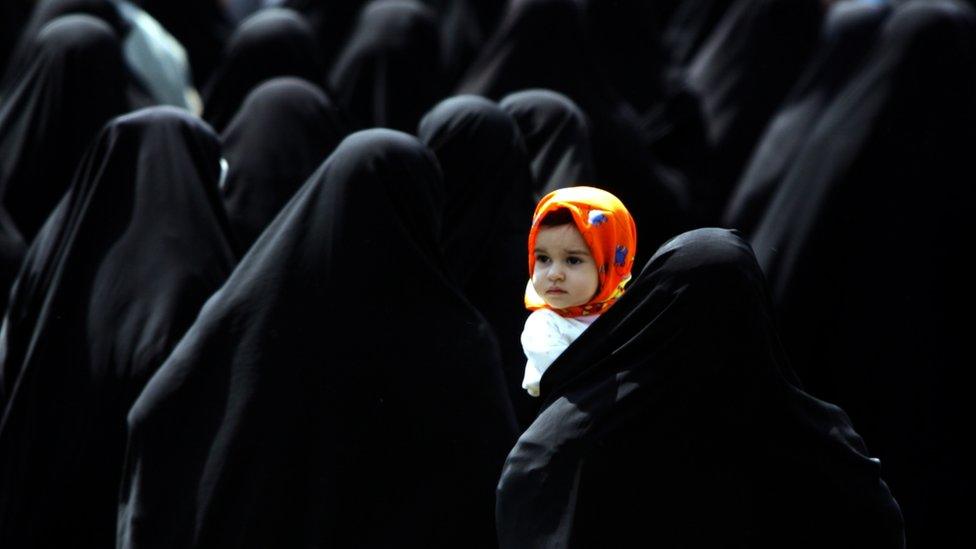 Dozens of women in black shadows with their back to the camera, expect for one little girl held in her mothers arms in a colourful headscarf in April 2006