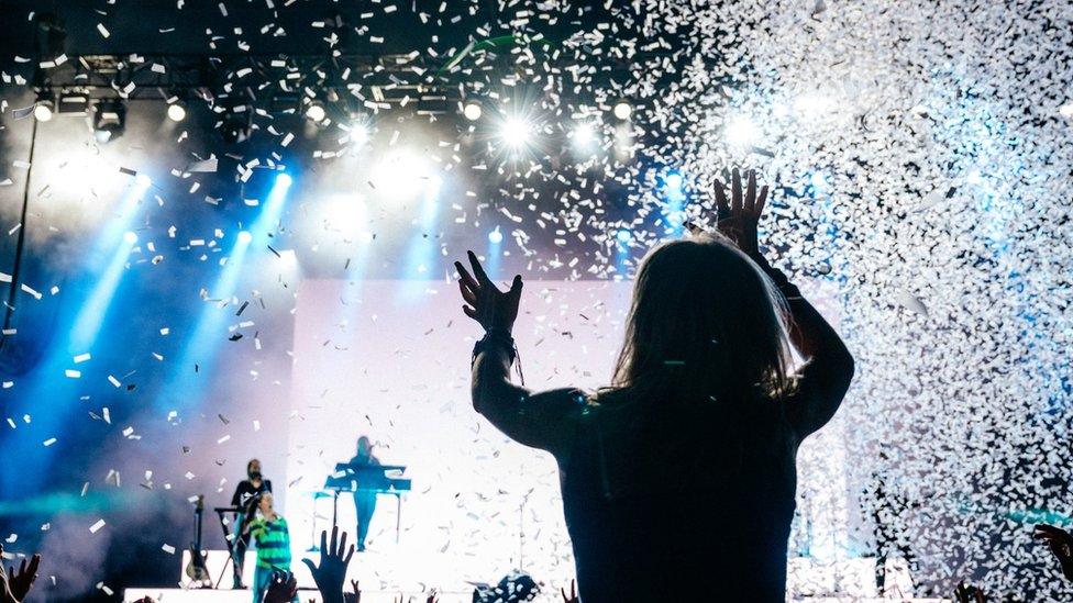 A girl enjoying a band playing at the Big Feastival as she sits on someone's shoulders and confetti falls