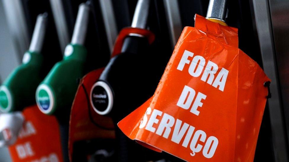 A close-up photograph shows fuel pump nozzles docked in their pumping station. One is wrapped in an orange sign reading "For de Servico" - the Portuguese for out of service