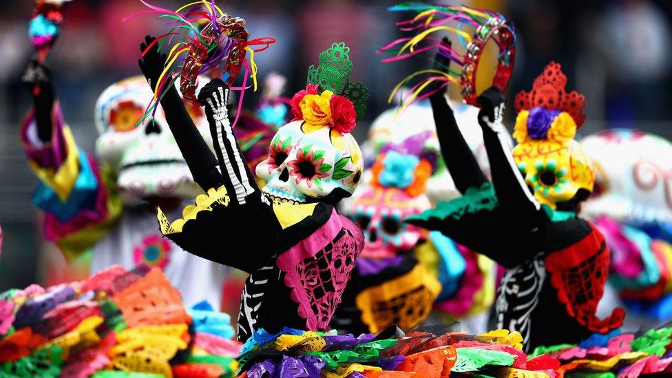 Dancers wearing pinks, yellows, blacks, reds and greens and large skulls dance with tambourines.