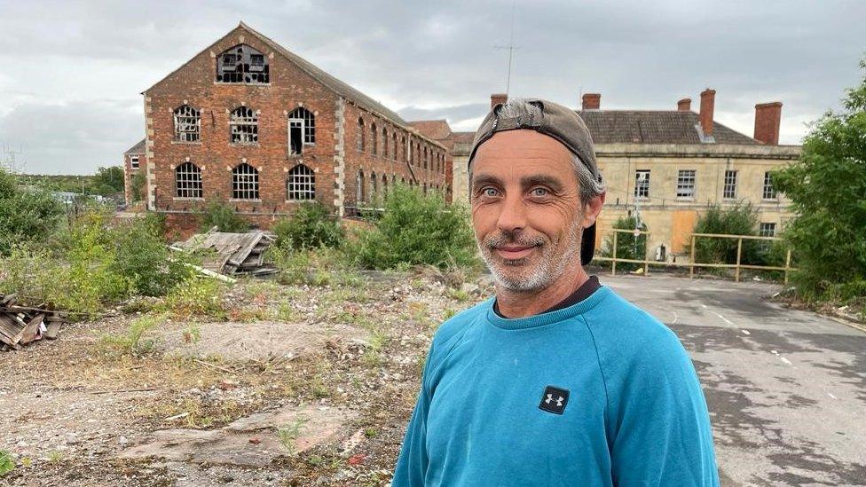 Builder Jon Kimber wearing a baseball cap back to front standing in front of the factory building formerly worked in the Bowyers factory