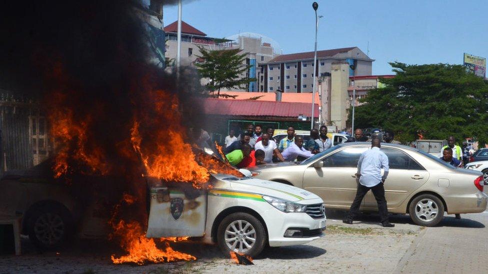 People stand next to a police car that has set alight