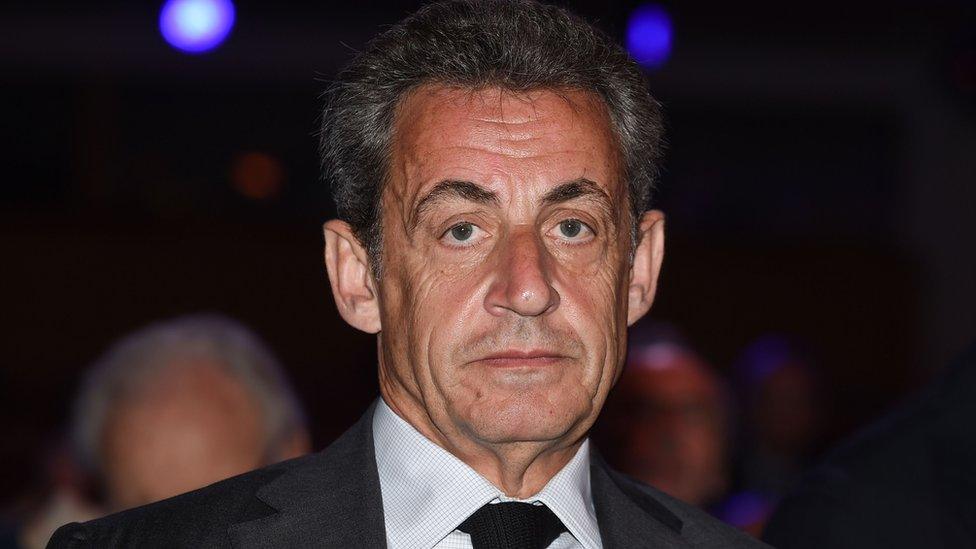 Nicolas Sarkozy, dressed in a dark suit and tie at night, looks directly into the camera in this close-up 2018 photo