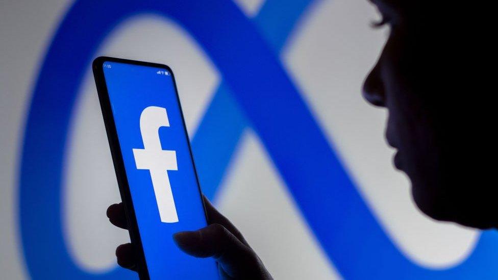 A woman holds a smartphone with Facebook's logo displayed against a background of Meta's logo