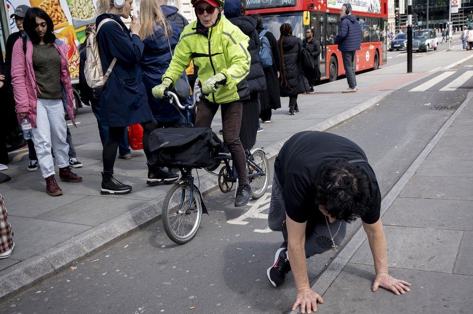 As a cyclist pedals past, a woman stands up again after sitting down in the cycle lane that's behind the bus stop shelter outside St Thomas' Hospital