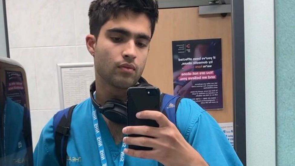 A man in medical scrubs and an NHS lanyard takes a selfie with a mobile phone in the mirror of a public toilet or changing room. He's wearing a pair of over-ear headphones around his neck and looking at the device as he takes the shot. Behind him a pine-coloured exit door with an information poster can be seen, and a cleaning logging form on the wall.