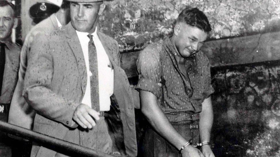 Black and white picture of a young man in handcuffs being led down a slope by a man in a Stetson hat