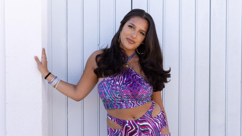 Priya leaning against a white wall, she is wearing her hair in loose curls and a purple top and trousers.