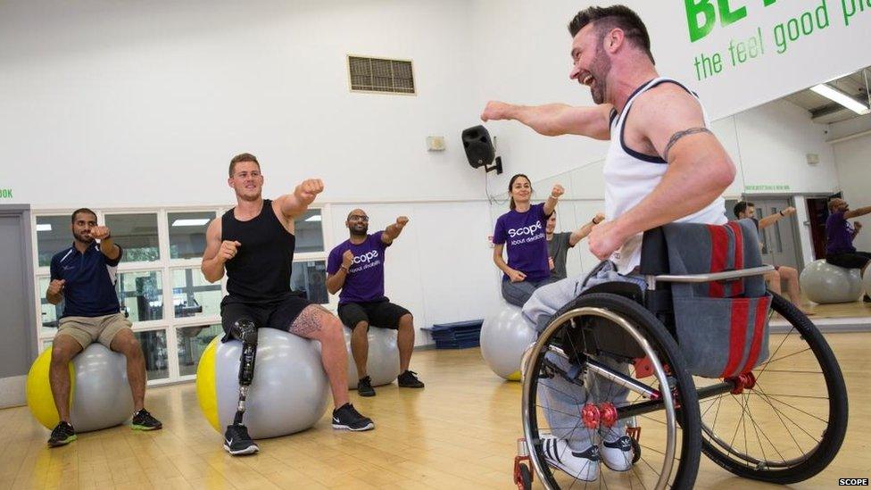 Kris teaching a gym class. Amputee model Jack Eyers is there and apart from Kris they are all doing a punching movement while sitting on gym balls